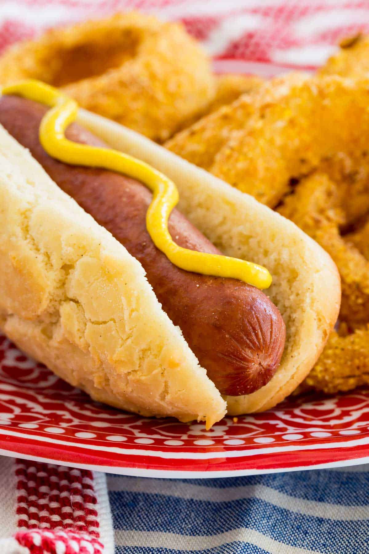 A gluten free hot dog topped with yellow mustard, served on a plate next to onion rings.