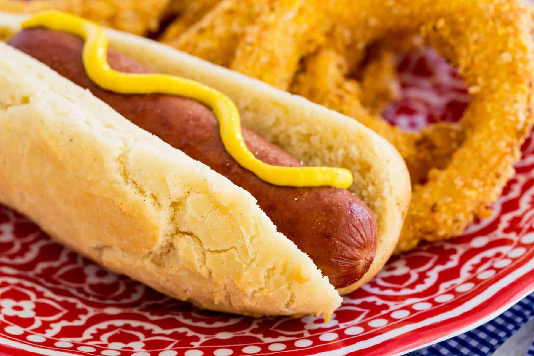 A gluten free hot dog topped with yellow mustard, served on a plate next to onion rings.