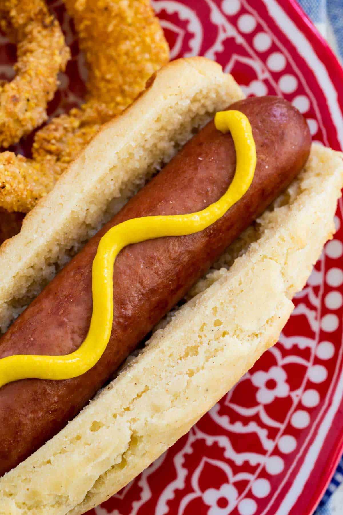 Top view of a gluten free hot dog topped with yellow mustard, served on a plate next to onion rings.