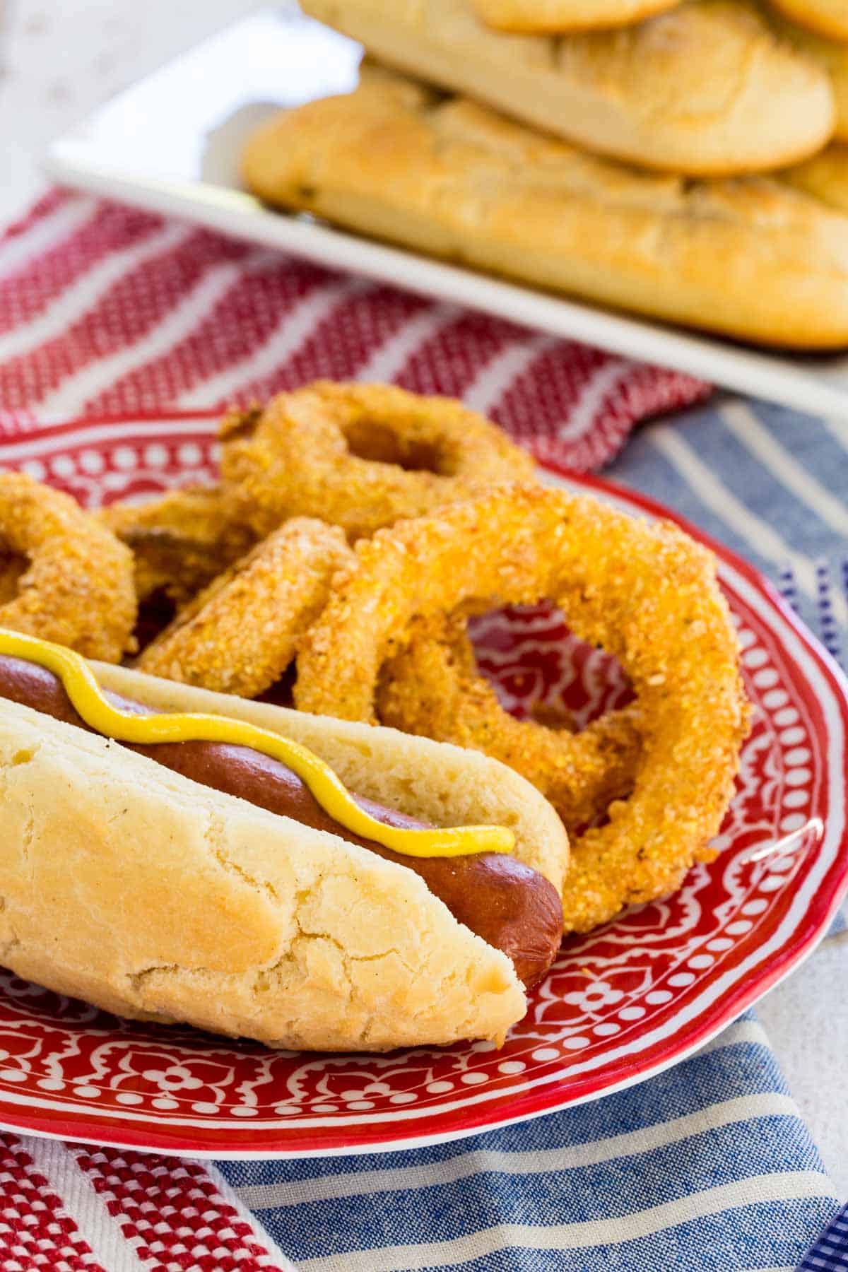 A gluten free hot dog topped with yellow mustard, served on a plate next to onion rings.
