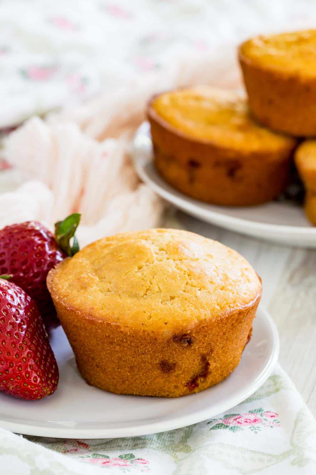A strawberry muffin on a white plate next to a strawberry, with another plate of muffins in the background.