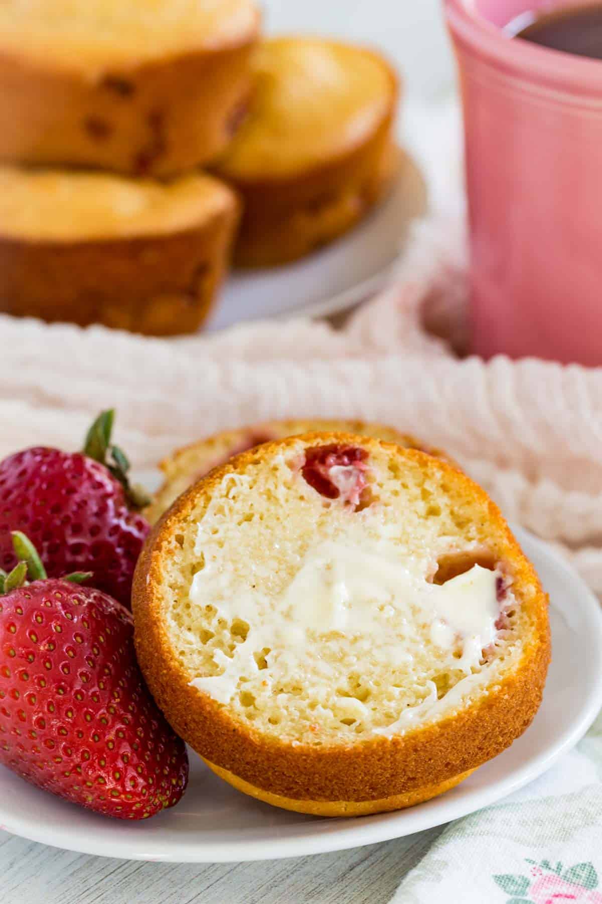 A halved strawberry muffin spread with butter on a plate next to a strawberry, with another plate of muffins and a cup of coffee in the background.