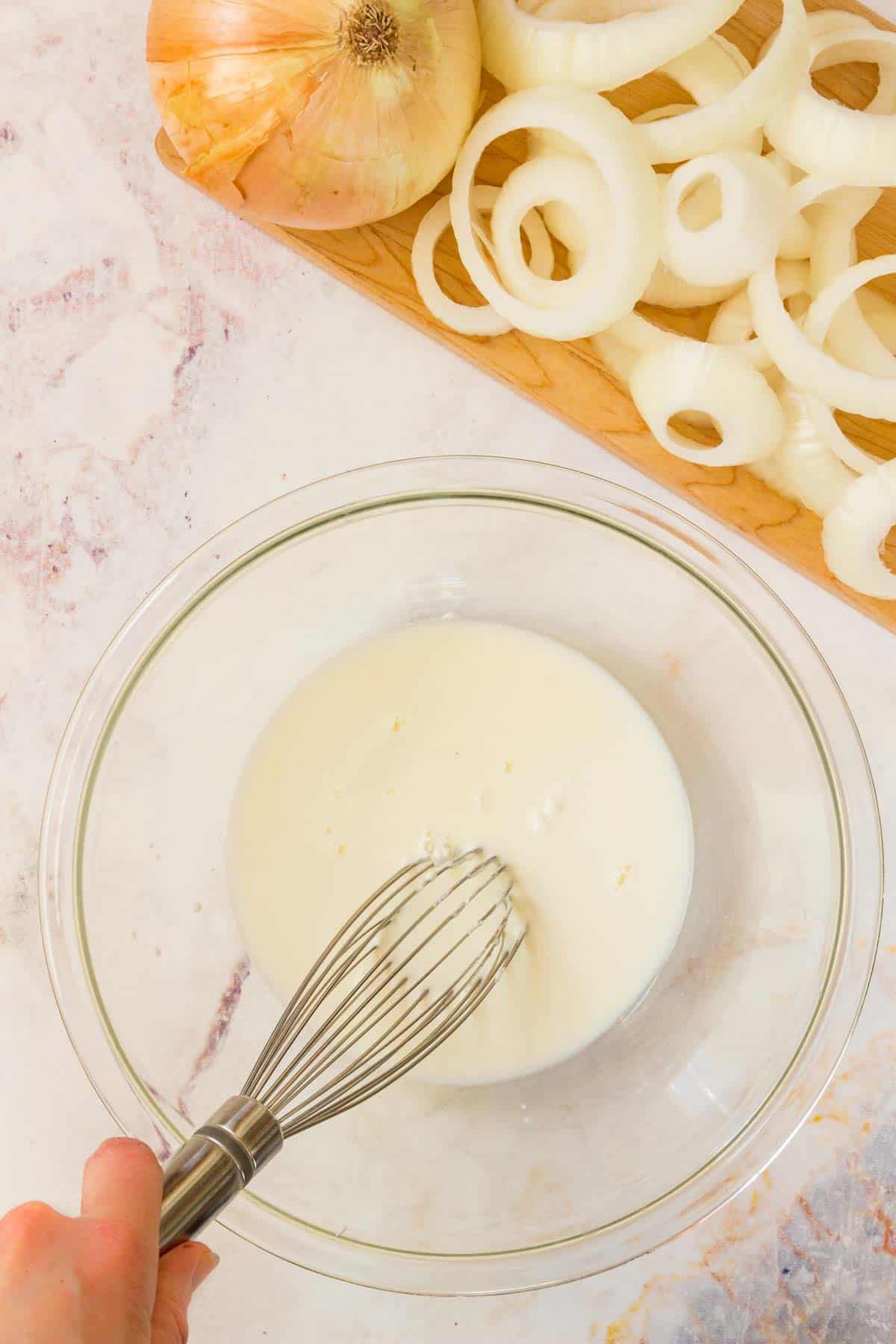 The milk coating ingredients are whisked together in a bowl next to a tray of sliced onions.