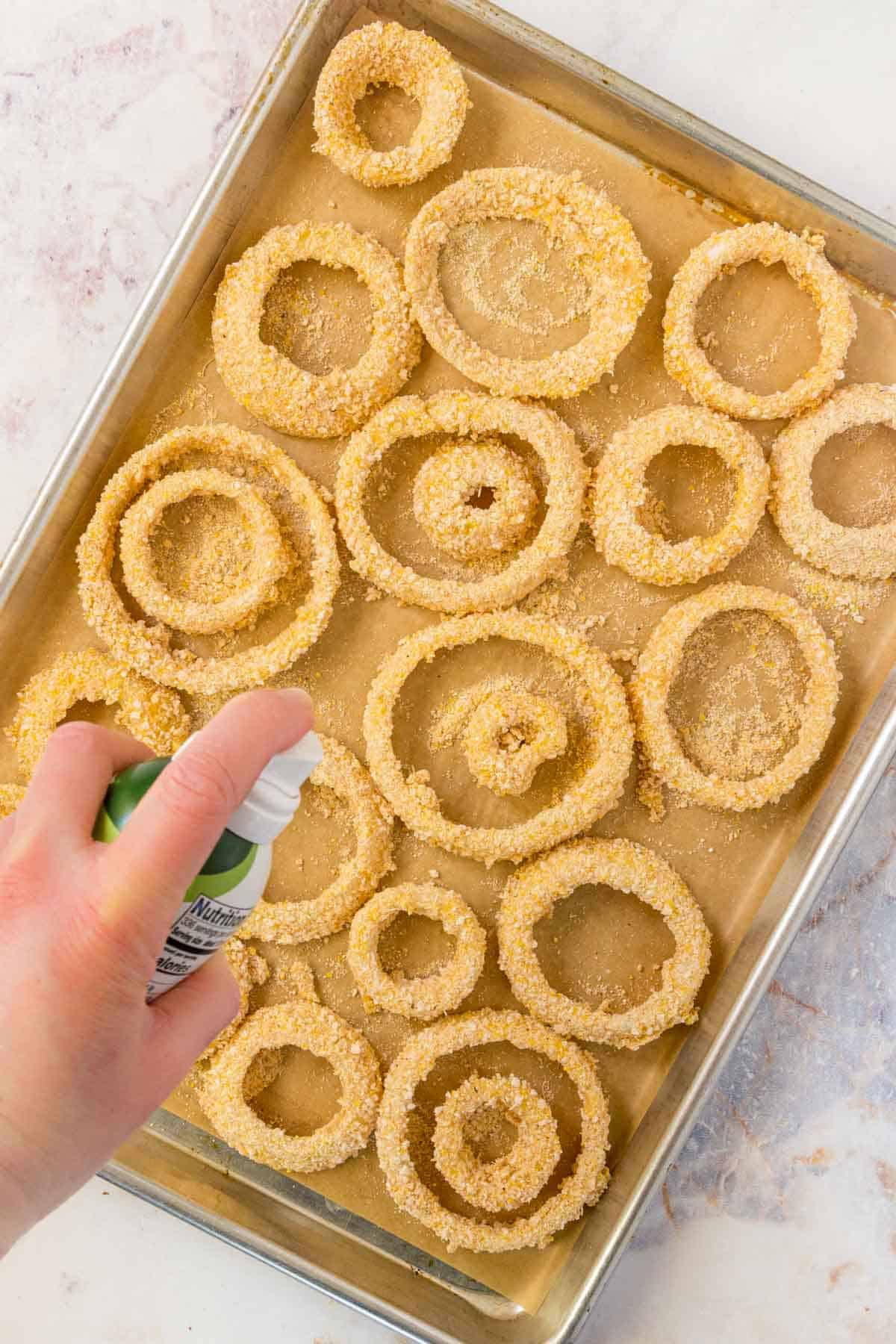 Breaded onions on a baking tray are sprayed with cooking oil prior to air frying.