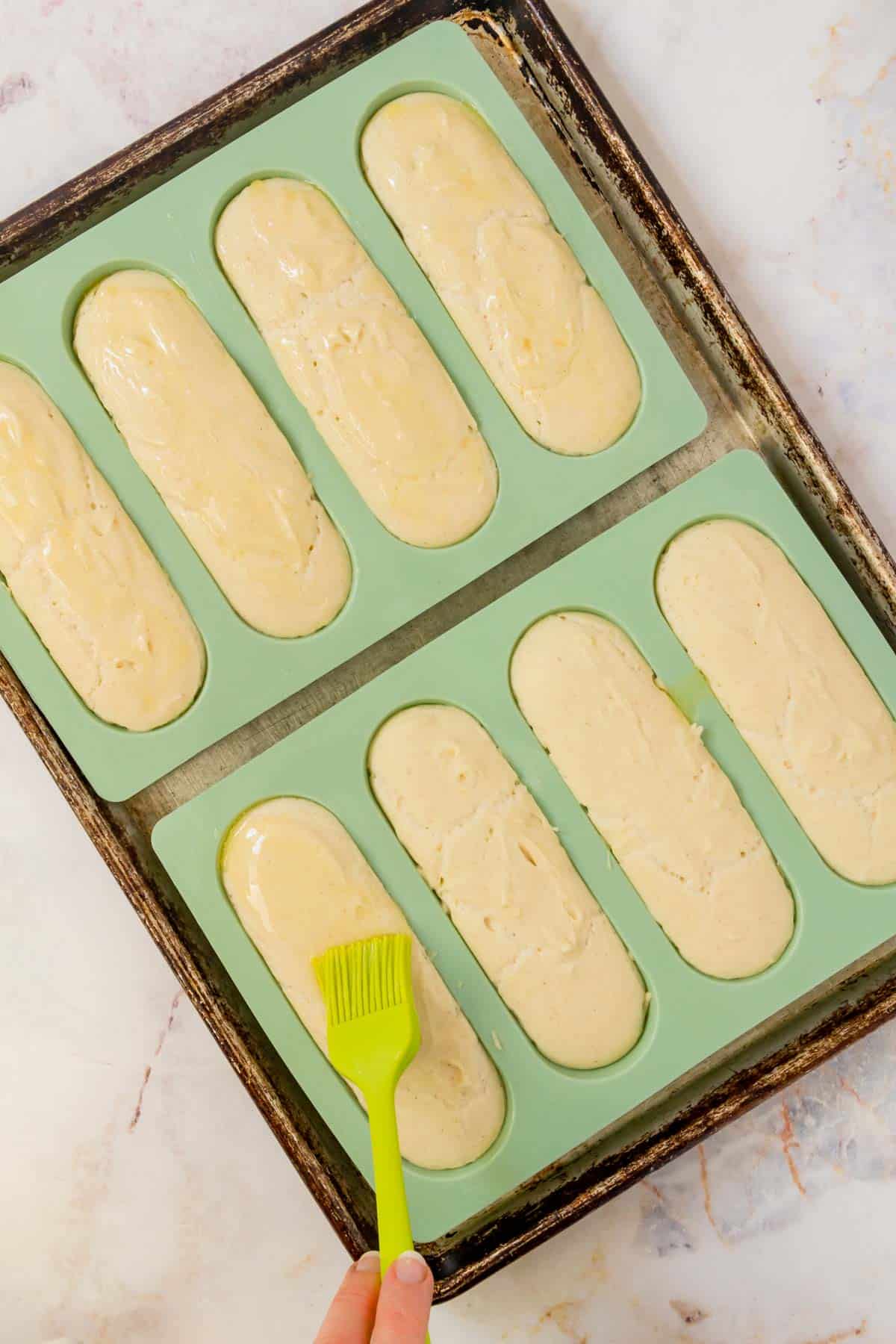 Butter is brushed overtop hot dog bun dough in a green silicone hot dog mold.