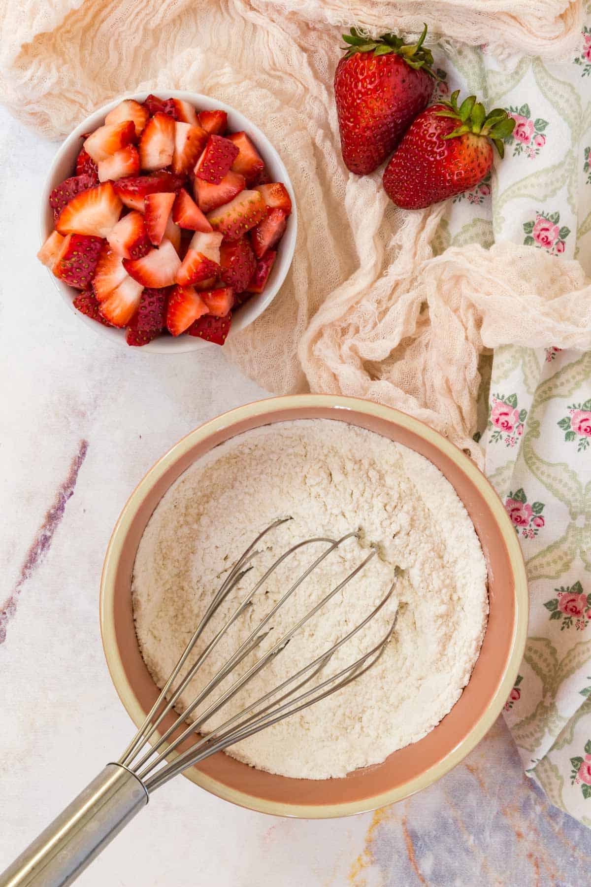 The dry ingredients for muffin batter are whisked together in a mixing bowl, next to a bowl of chopped strawberries.