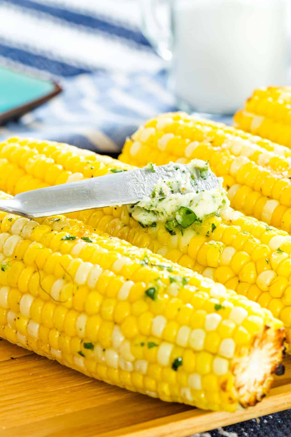 Close up of herb butter being spread over an ear of roasted corn on the cob, next to more ears of corn.
