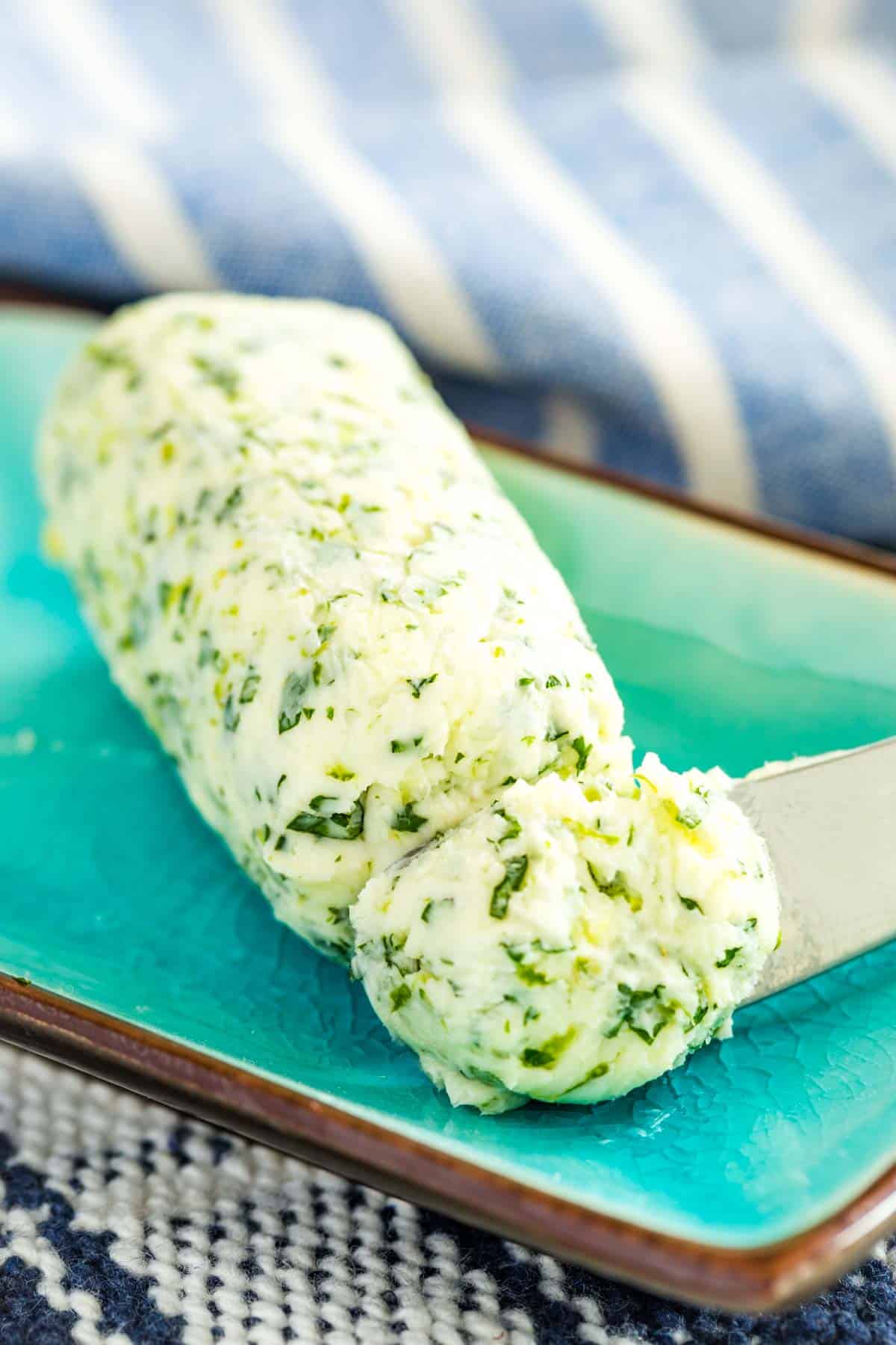 A log of herb butter on a turquoise blue plate, with a butter knife scooping from the end.