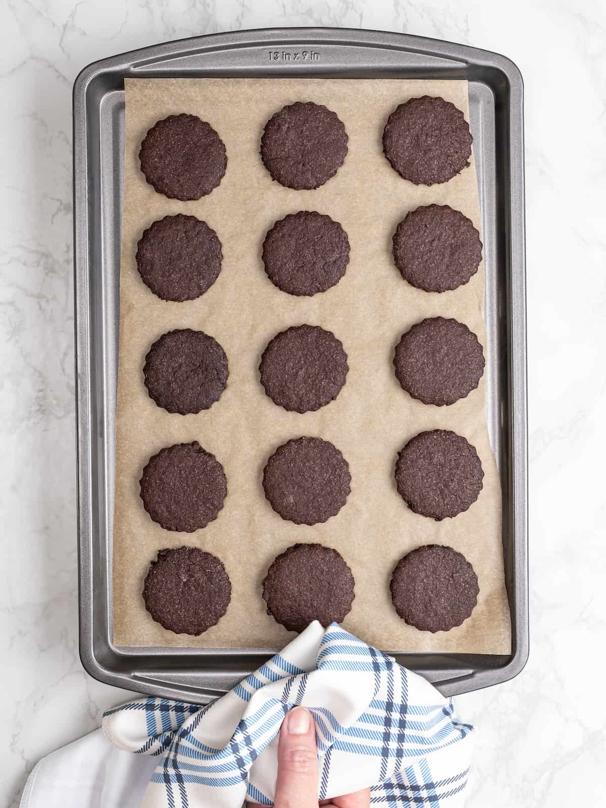 A hand holding a baking sheet filled with three rows of chocolate cookies.