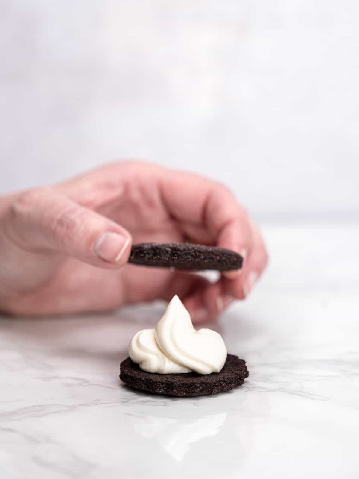 A hand places a chocolate cookie over a second cookie topped with vanilla cream, to form a sandwich.