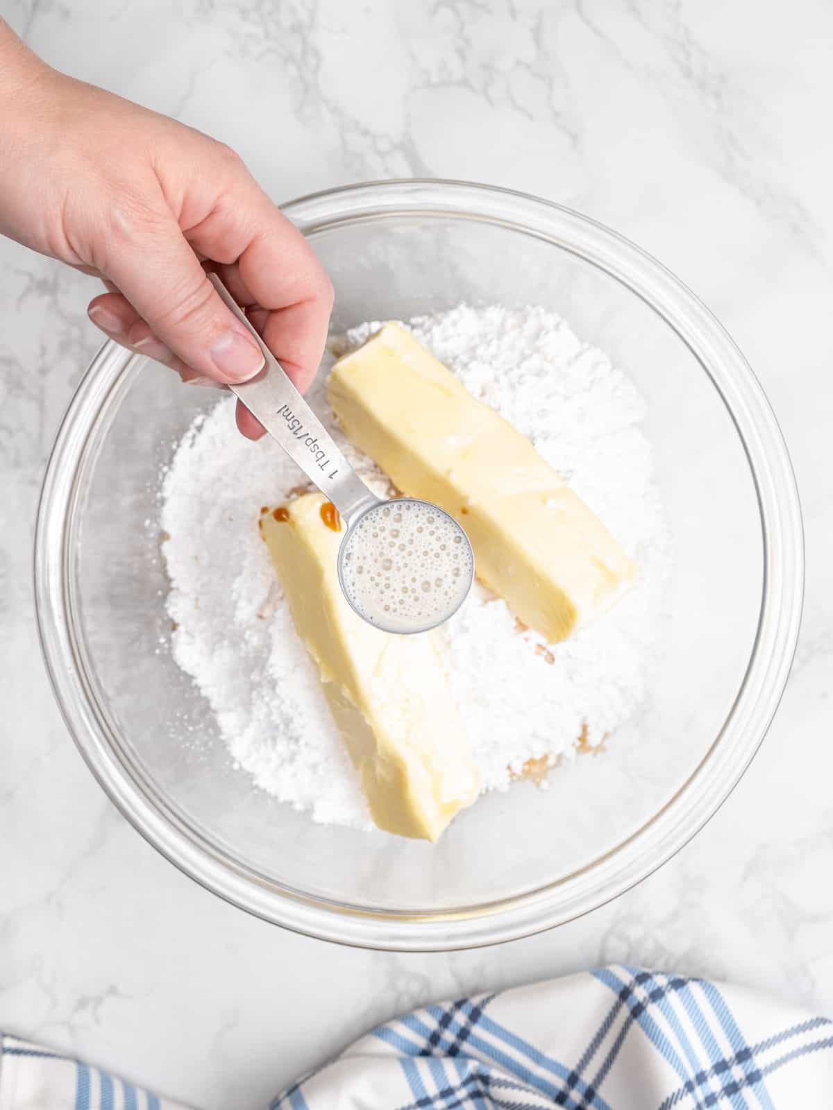 A hand holds a tablespoon of oat milk over a mixing bowl with powdered sugar and two butter sticks.