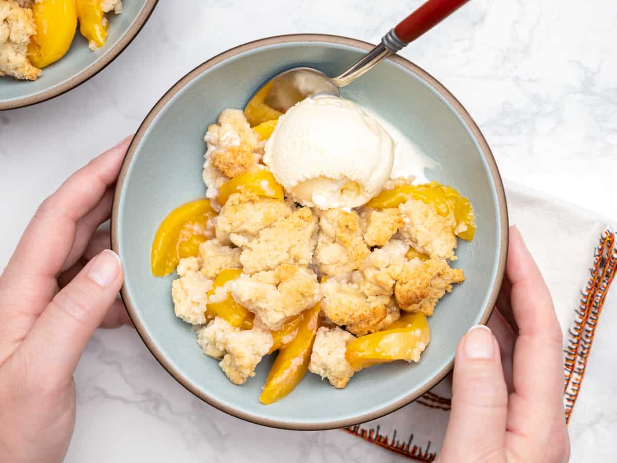 Gluten free peach cobbler topped with vanilla ice cream, in a bowl held by two hands.