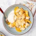 A bowl of gluten free peach cobbler topped with vanilla ice cream, with a spoon.