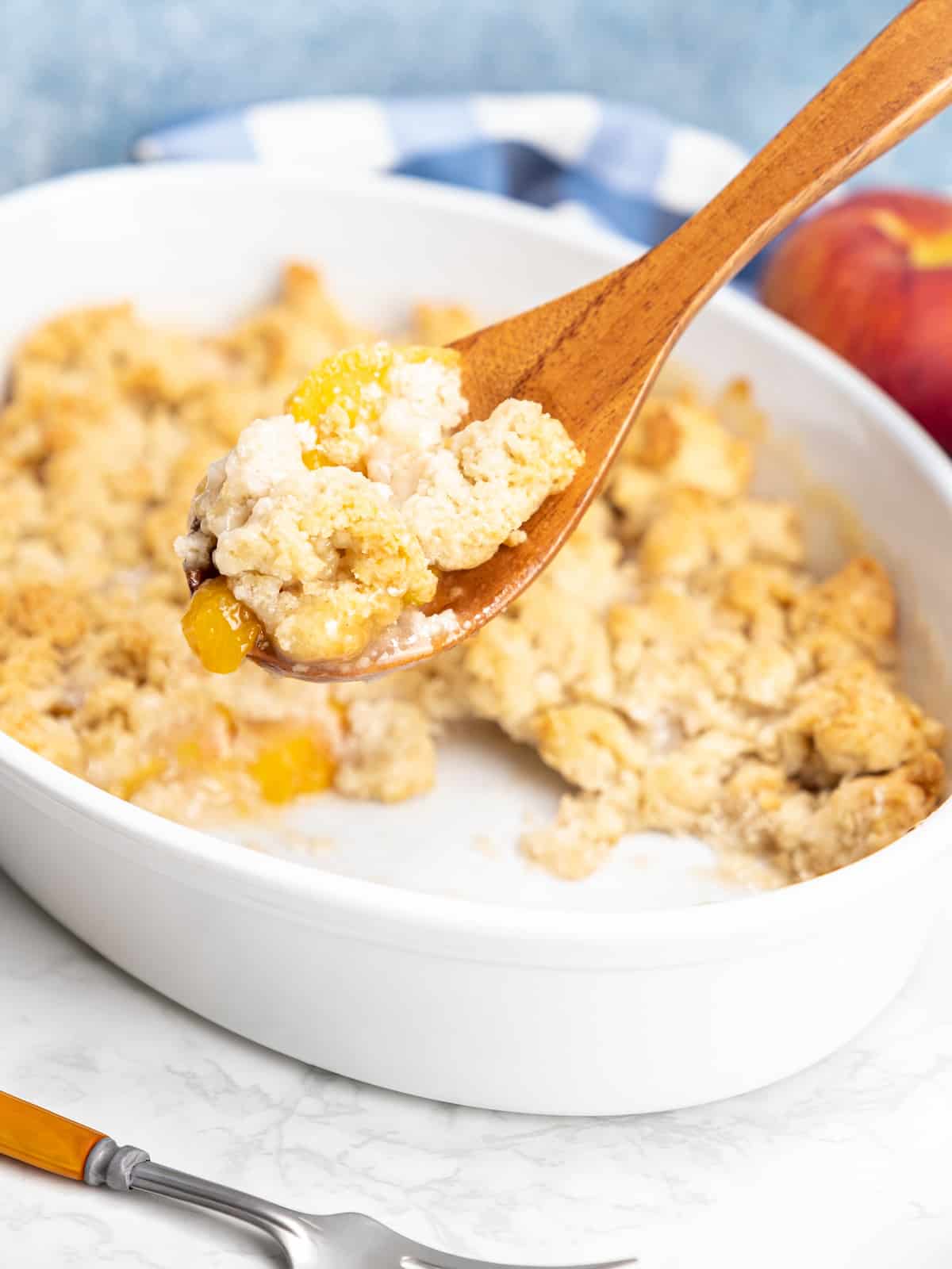 A wooden spoon lifts a serving of peach cobbler out of an oval baking dish.