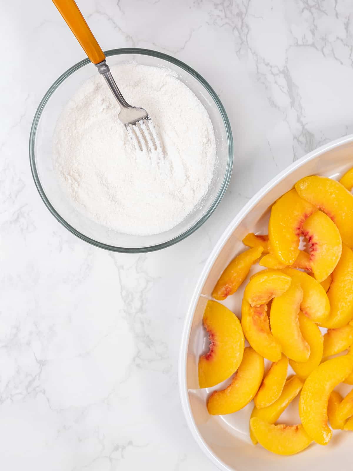 Sliced peaches in an oval baking dish, next to a bowl of sugar and flour whisked together with a fork.