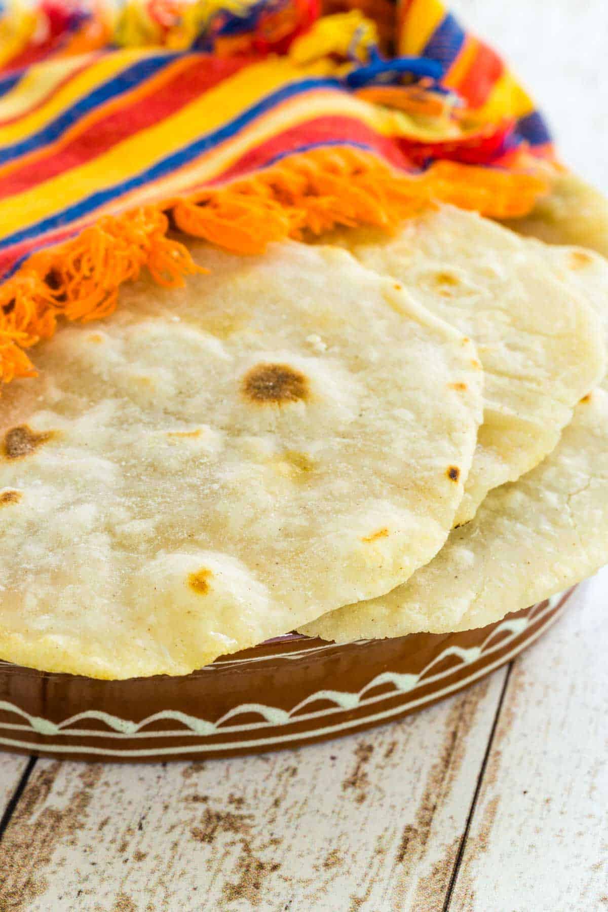 Gluten free tortillas stacked in a bowl beneath a colorful striped cloth to keep warm.