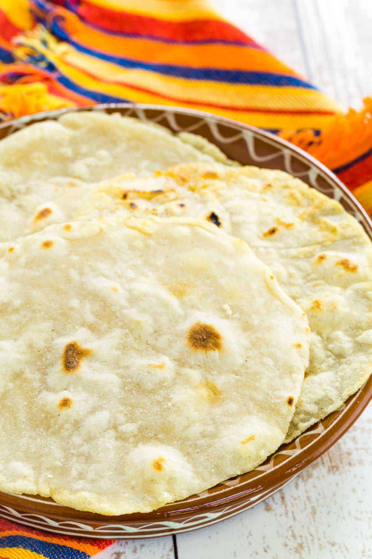 Gluten free tortillas stacked in a brown bowl with a colorful striped cloth in the background.