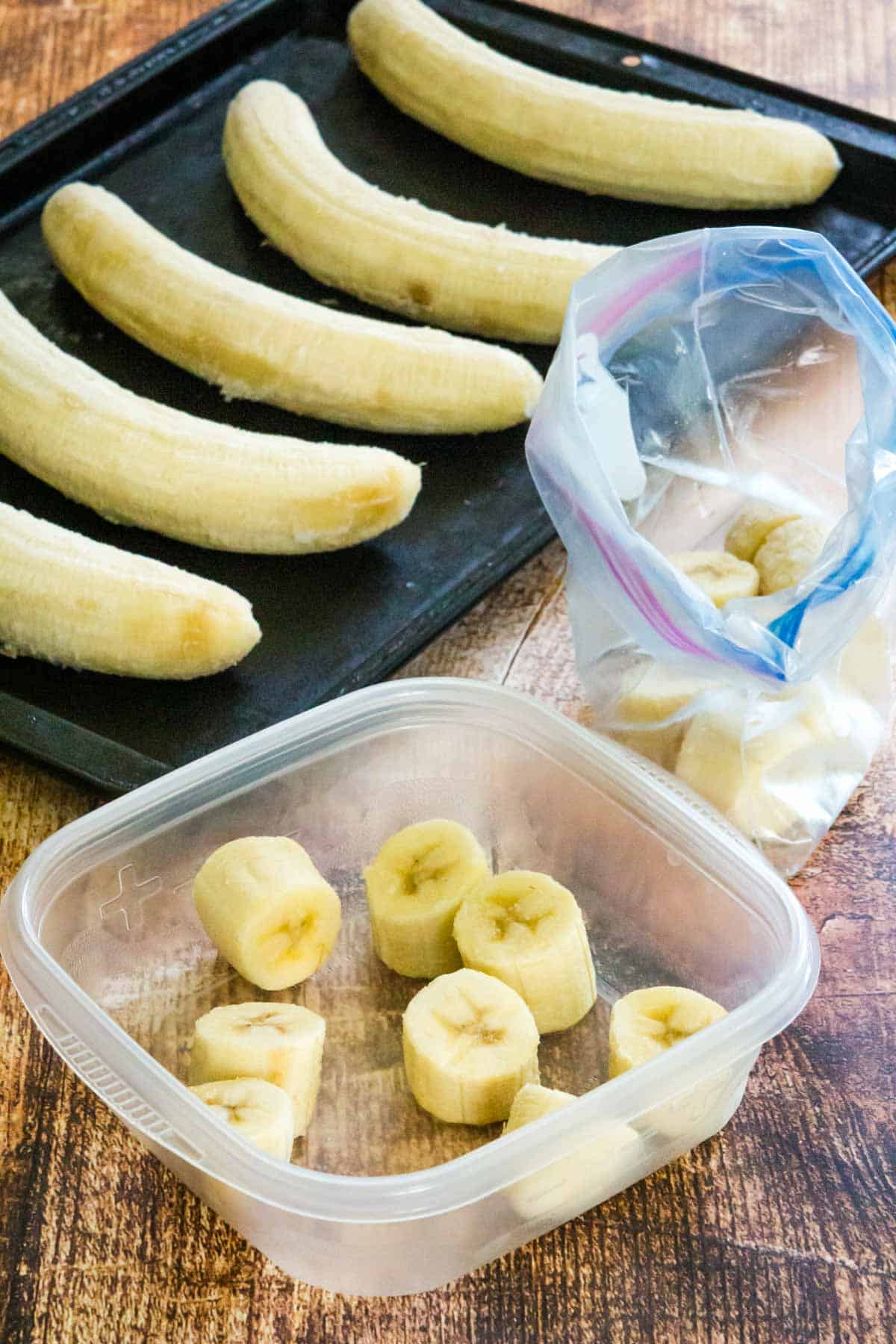 A baggie and a tupperware full of banana slices next to a baking sheet with five whole peeled bananas spread across it