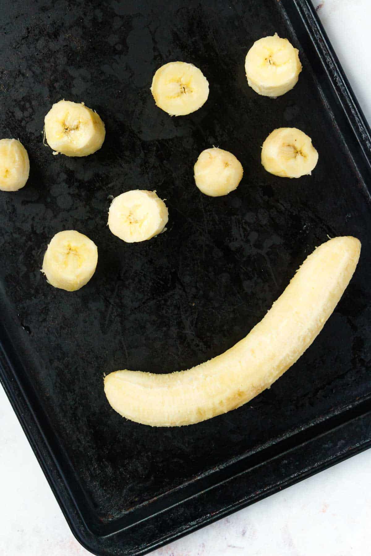 A whole banana next to eight banana slices spread out evenly on a baking sheet