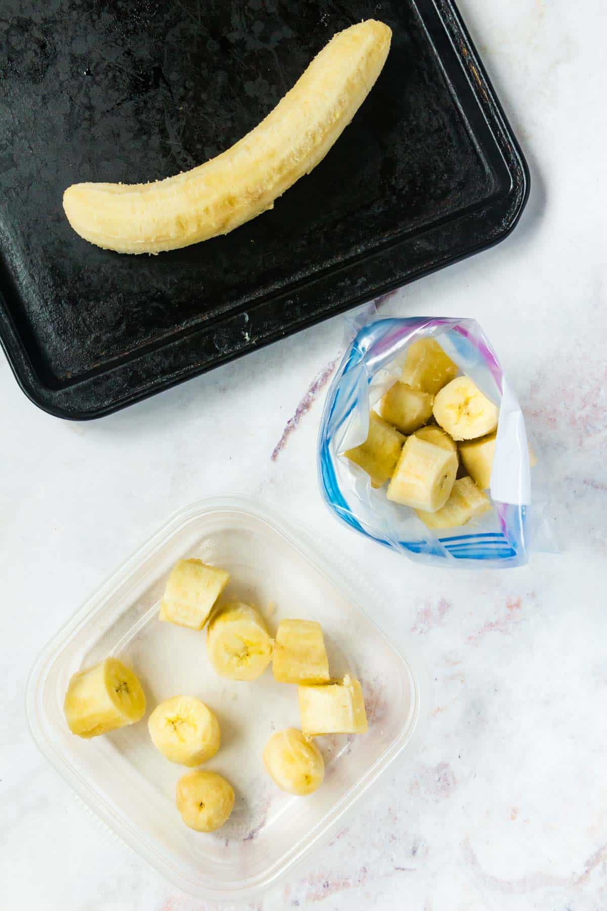 A frozen whole banana on a sheet pan, and chunks in a plastic bag and a plastic container.