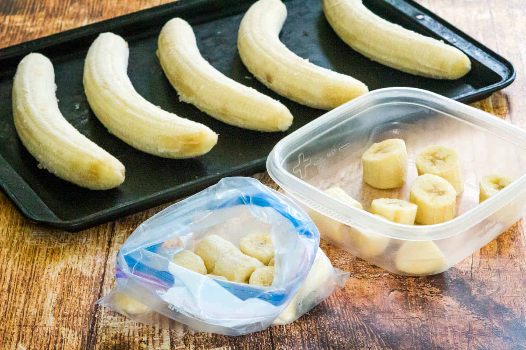 Five bananas without the peels lined up on a baking sheet next to a plastic container and a plastic baggie filled with sliced banana