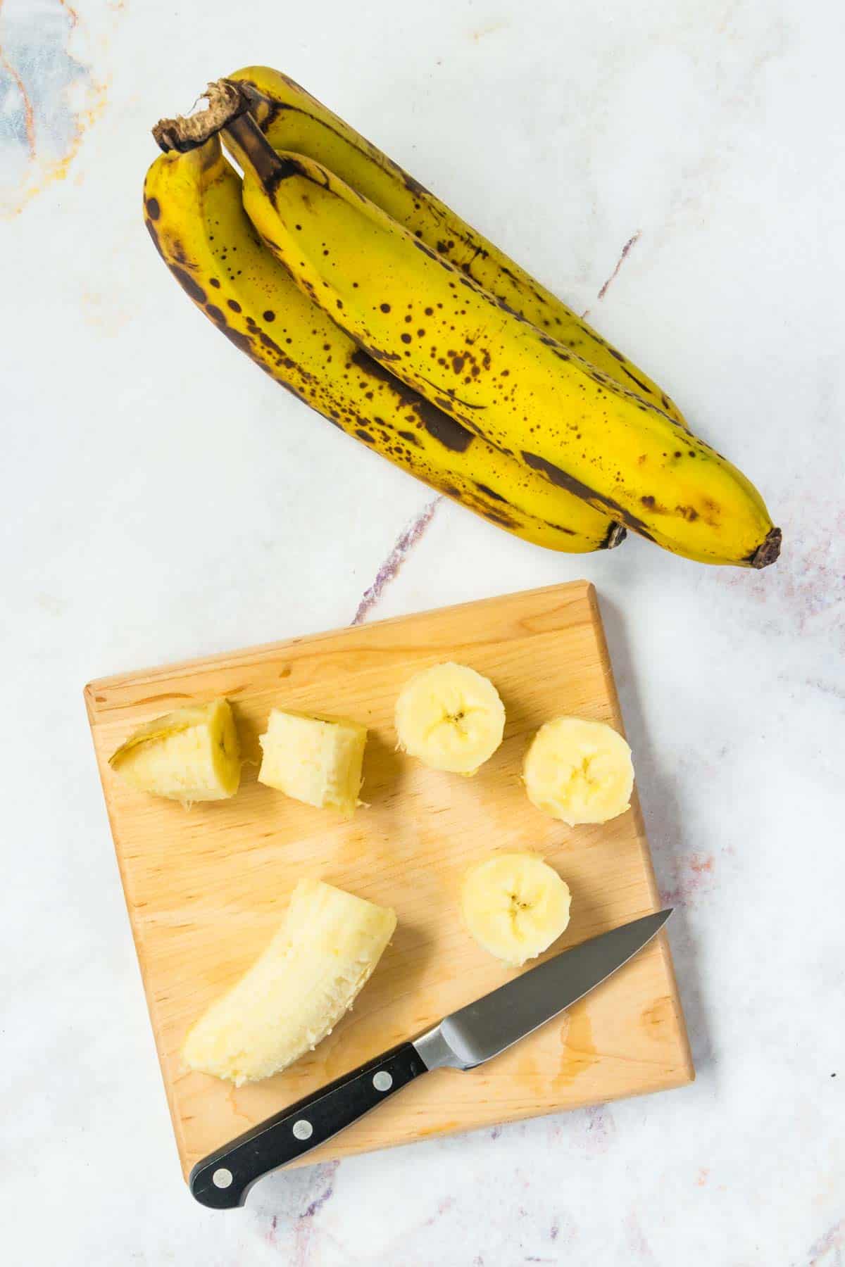 A bunch of bananas next to a cutting board where another banana is being sliced