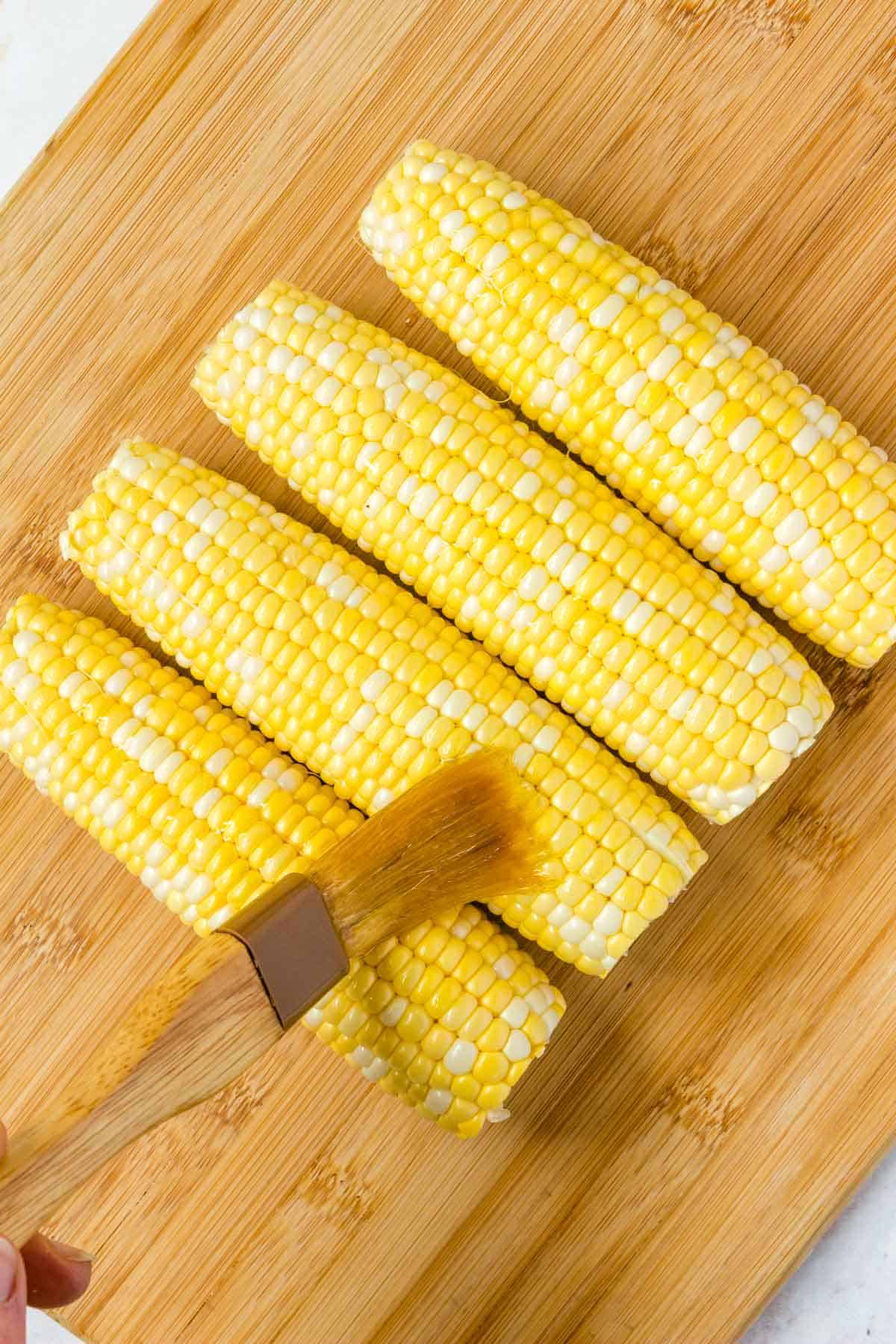 Top view of four ears of corn on a wooden cutting board, being brushed with olive oil.