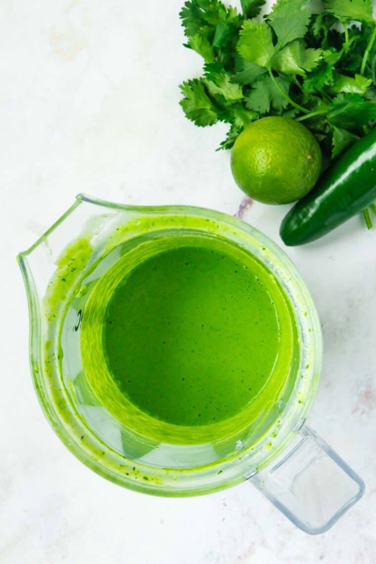 Blended Aji sauce in a blender jar next to a lime, a jalapeno, and some cilantro on a countertop.