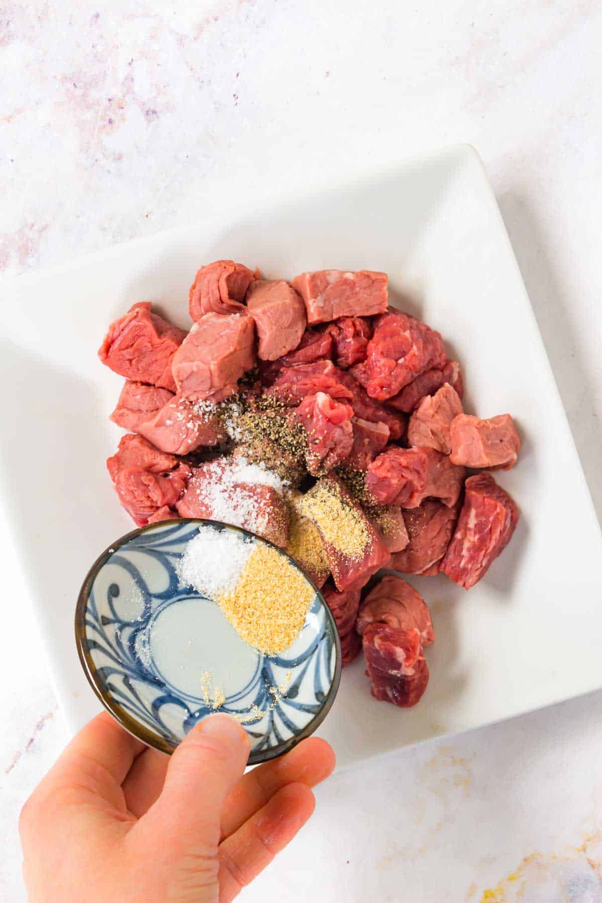 Sprinkling seasonings over steak cubes on a plate.