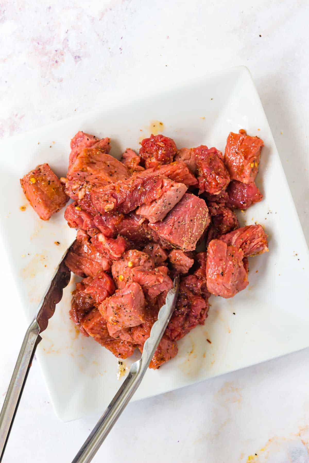 Using tongs to toss the seasoned steak cubes to coat them in honey and chili paste.