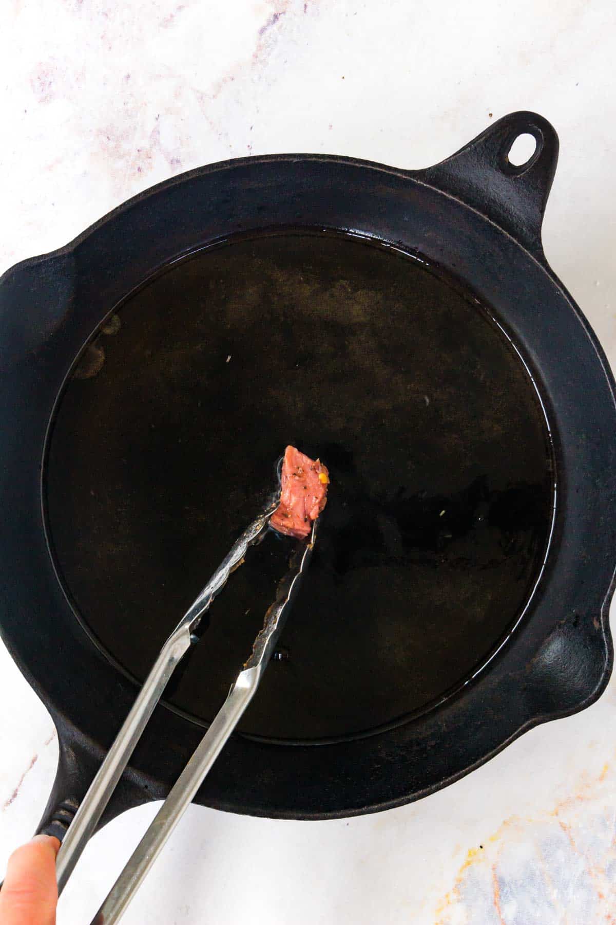 Tongs placing one steak cube into a cast iron skillet.