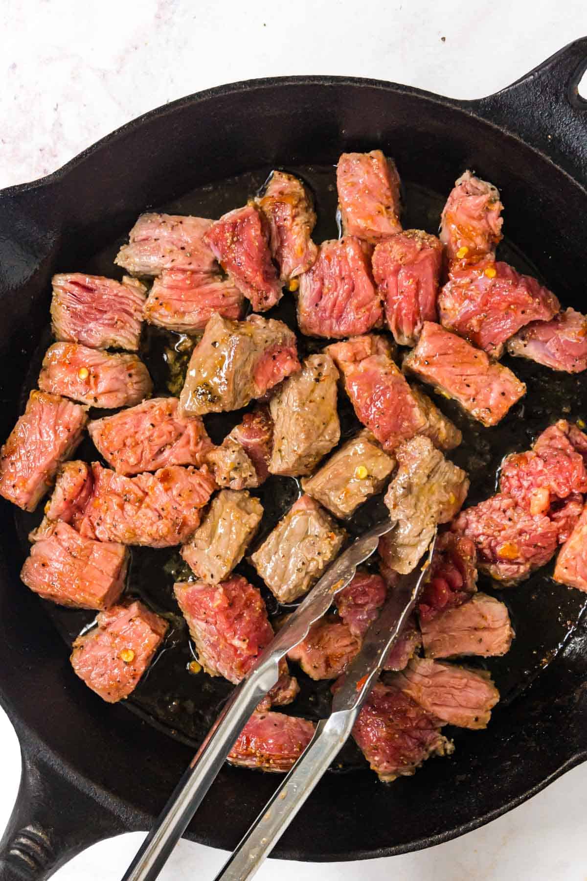 Flipping the steak bites in the cast iron skillet with tongs.