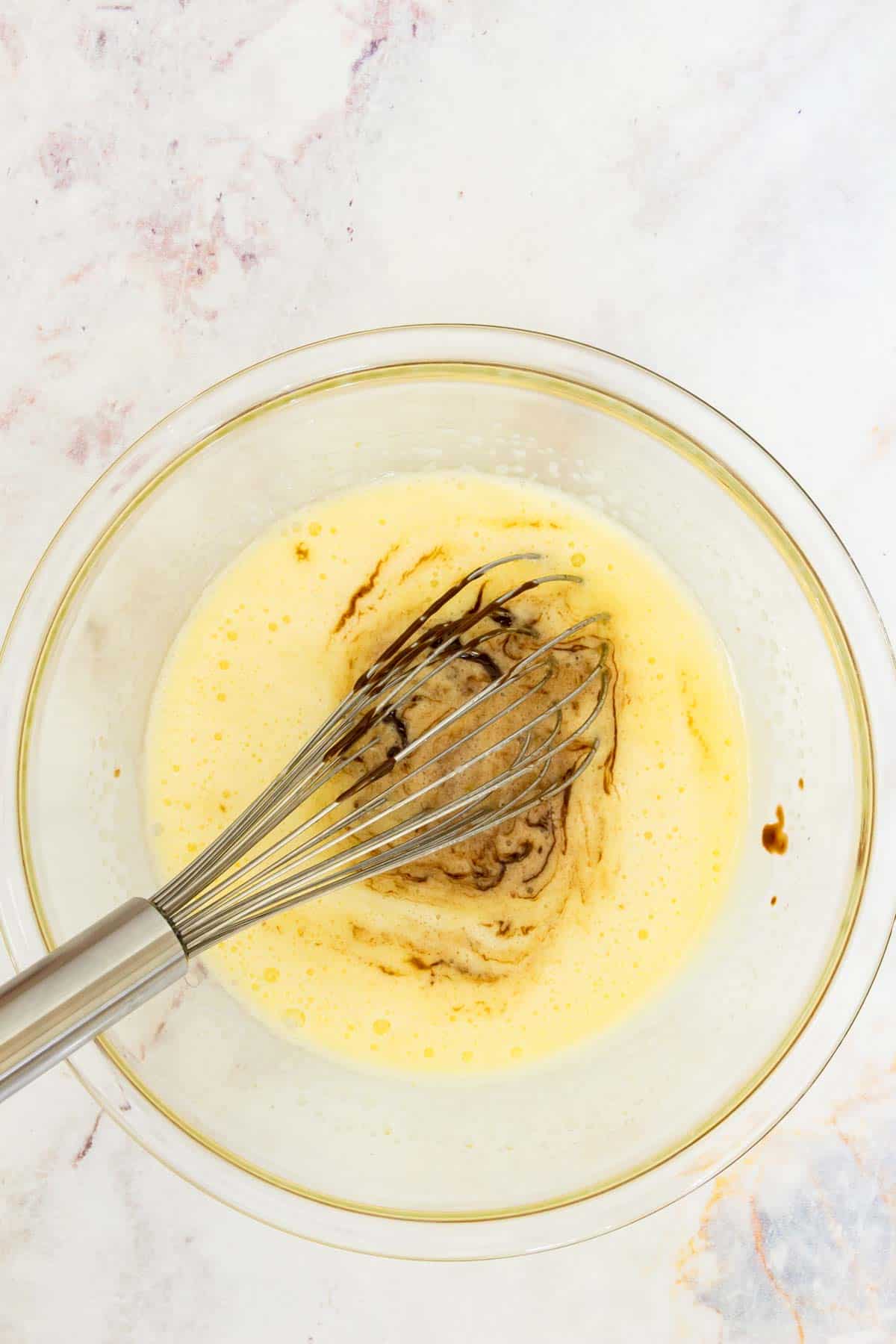 Melted chocolate is whisked into an egg and sugar mixture in a clear mixing bowl.