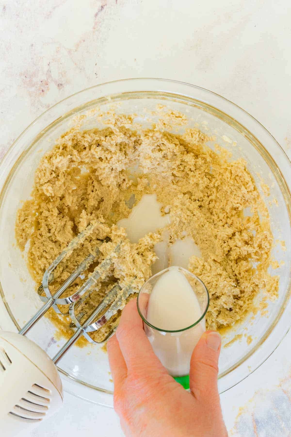 Milk is poured into cookie dough in a mixing bowl, next to a hand mixer.