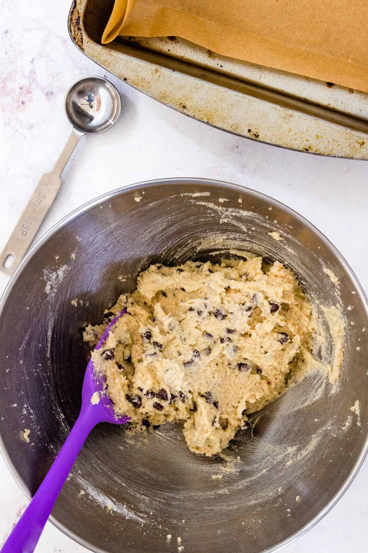 Gluten free edible chocolate chip cookie dough comes together in a metal mixing bowl with a purple spoon.