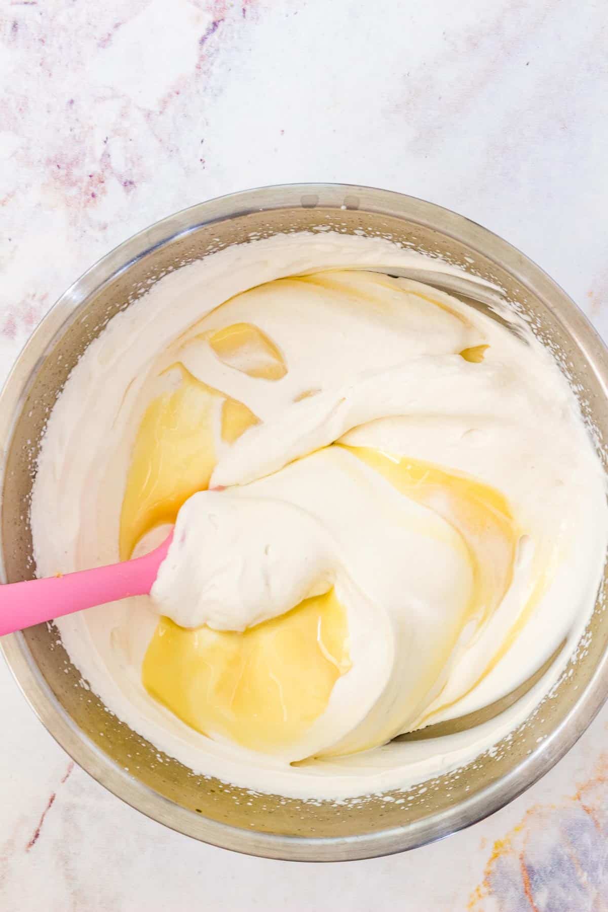 Sweetened condensed milk is folded into whipped cream with a pink stirring spoon.