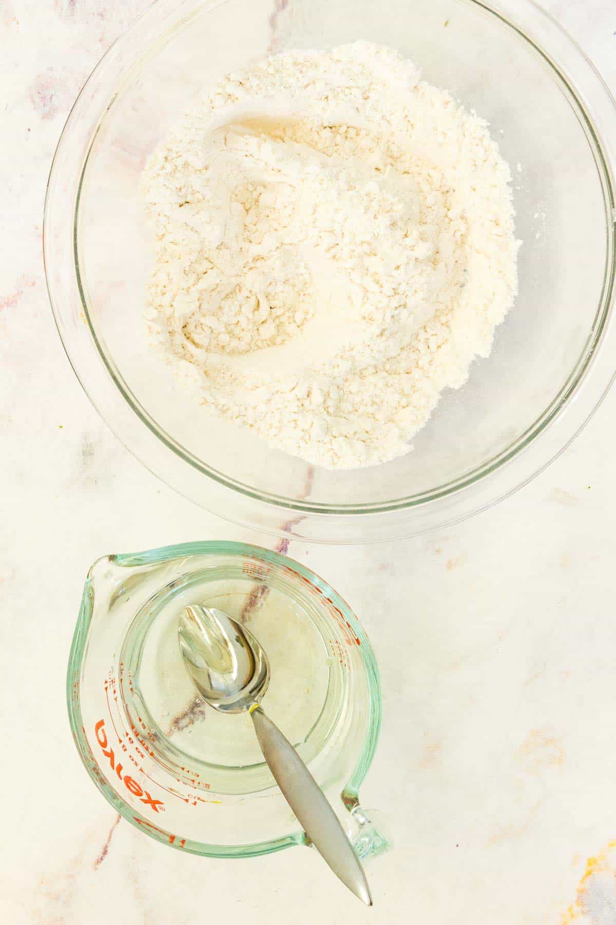 The flour and dry ingredients in a mixing bowl next to a measuring cup of warm water and a spoon.