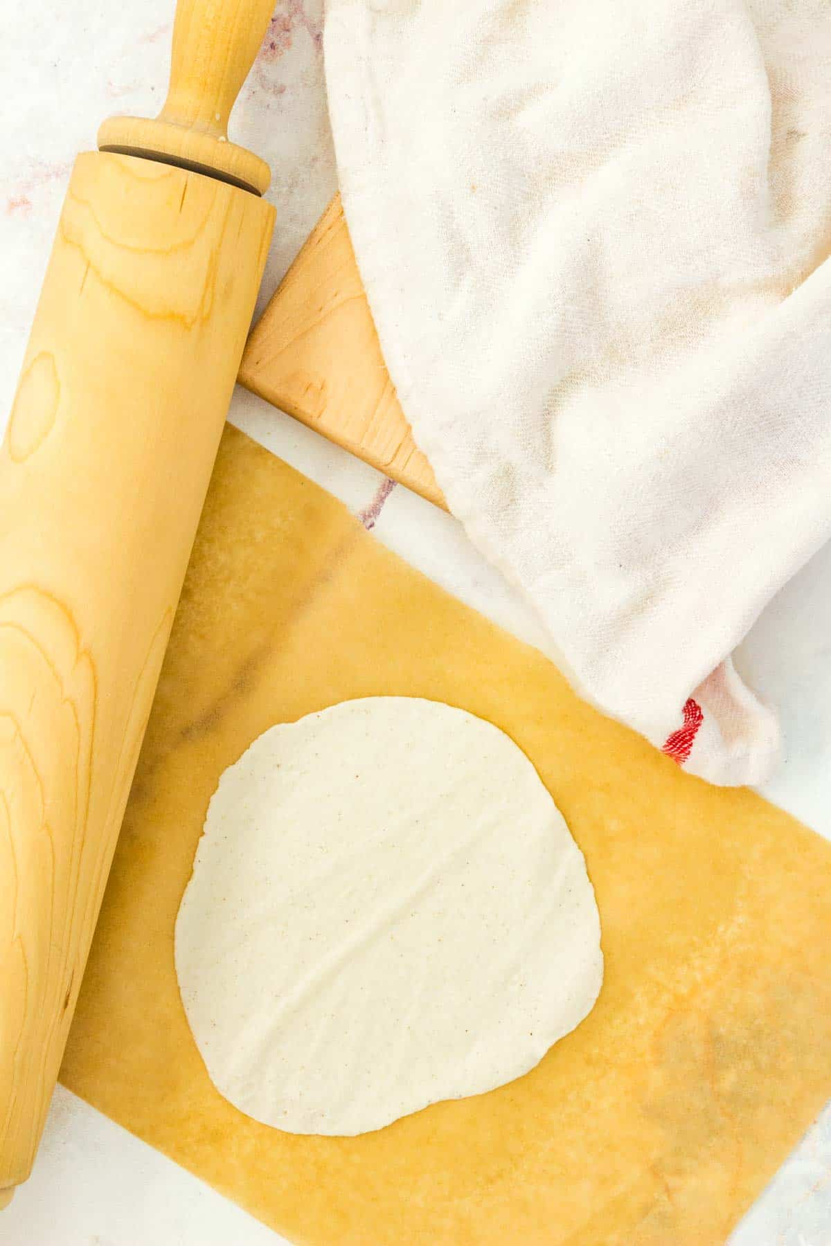 A flattened gluten free tortilla on a wooden board, next to a rolling pin and a white cloth.