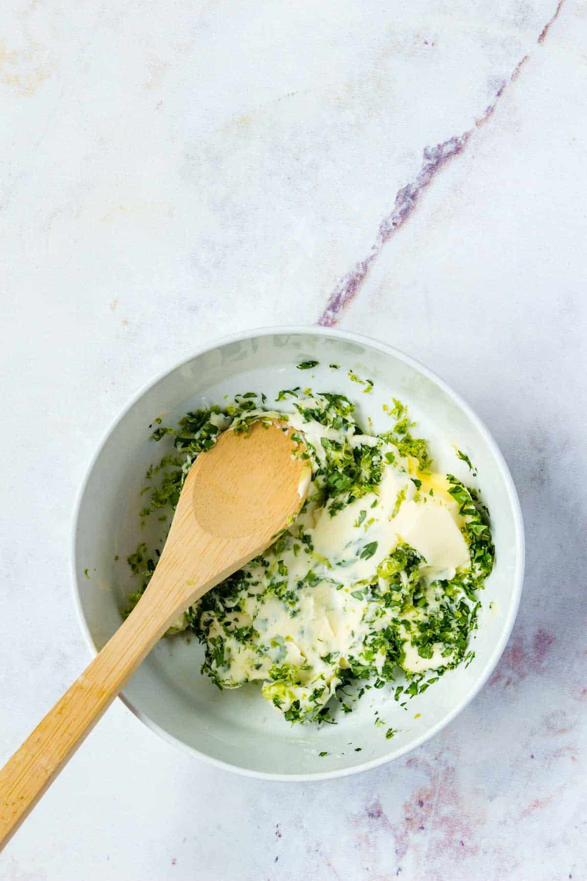 Fresh herbs are pressed into butter in a bowl with a wooden spoon.