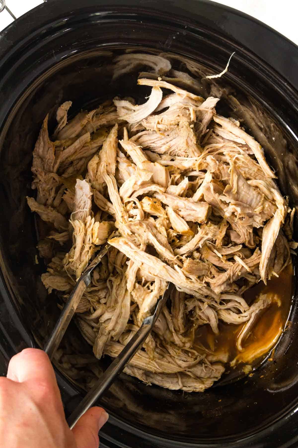 Using tongs to toss thw shredded pork in the juices in the crockpot.