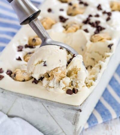 An ice cream scoop serves chocolate chip cookie dough no-churn ice cream from a metal loaf pan, topped with chunks of cookie dough and chocolate chips.
