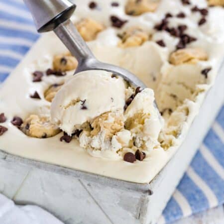 An ice cream scoop serves chocolate chip cookie dough no-churn ice cream from a metal loaf pan, topped with chunks of cookie dough and chocolate chips.