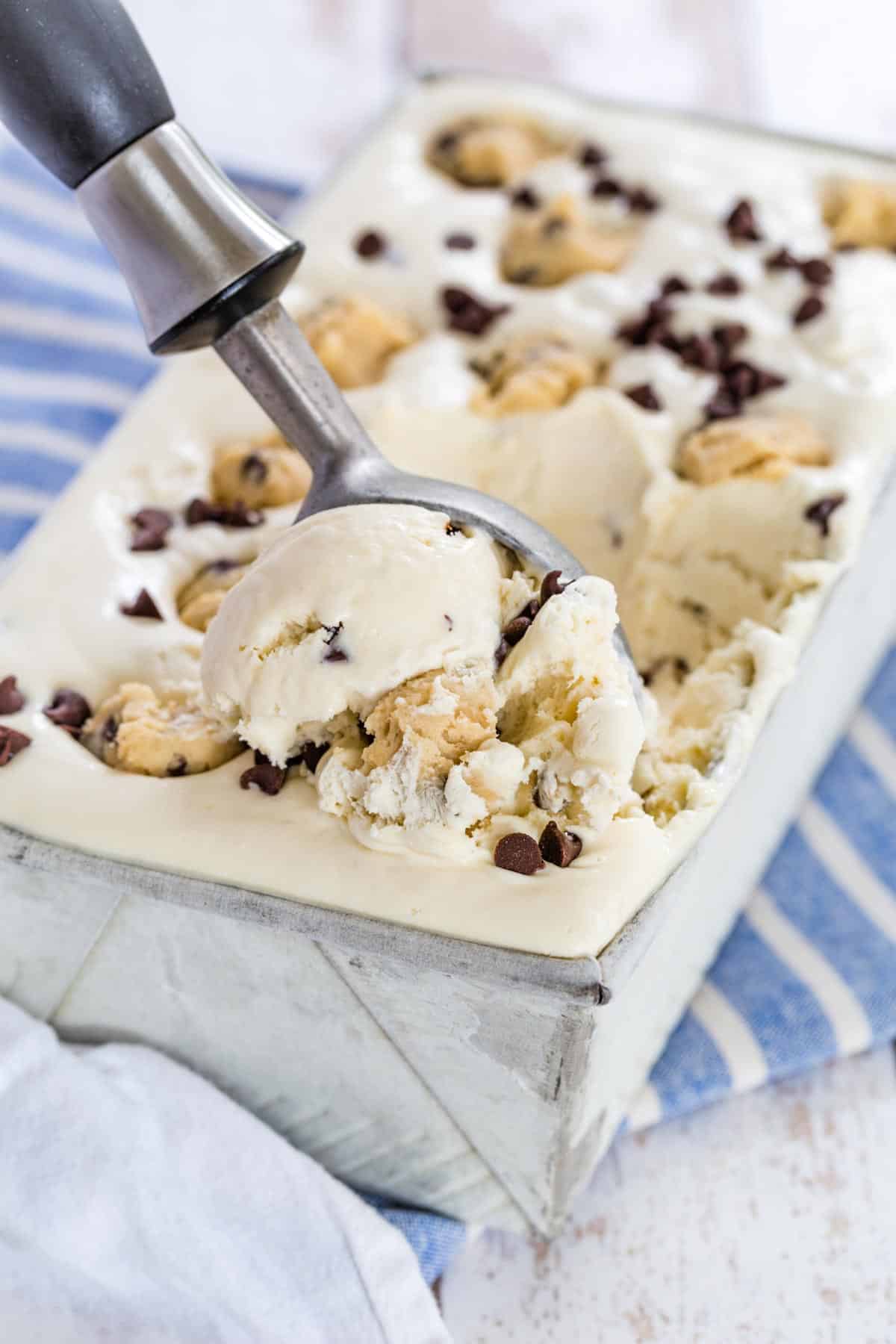An ice cream scoop serves chocolate chip cookie dough no-churn ice cream from a metal loaf pan, topped with chunks of cookie dough and chocolate chips.