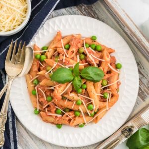 A bowl of penne pasta with tomato cream sauce and peas, and garnished with basil and parmesan cheese on a wooden tray.
