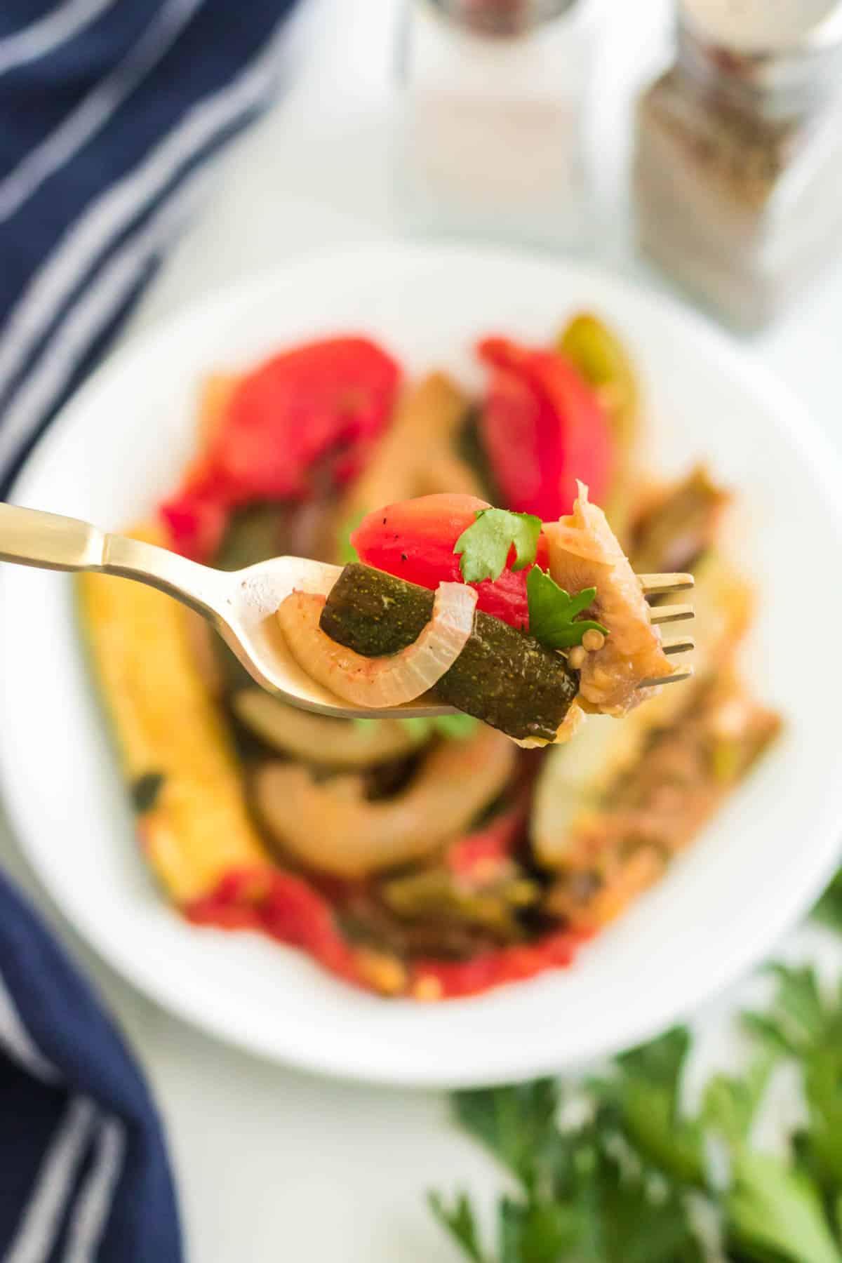A bite of ratatouille on a fork with a plate of vegetable casserole in the background