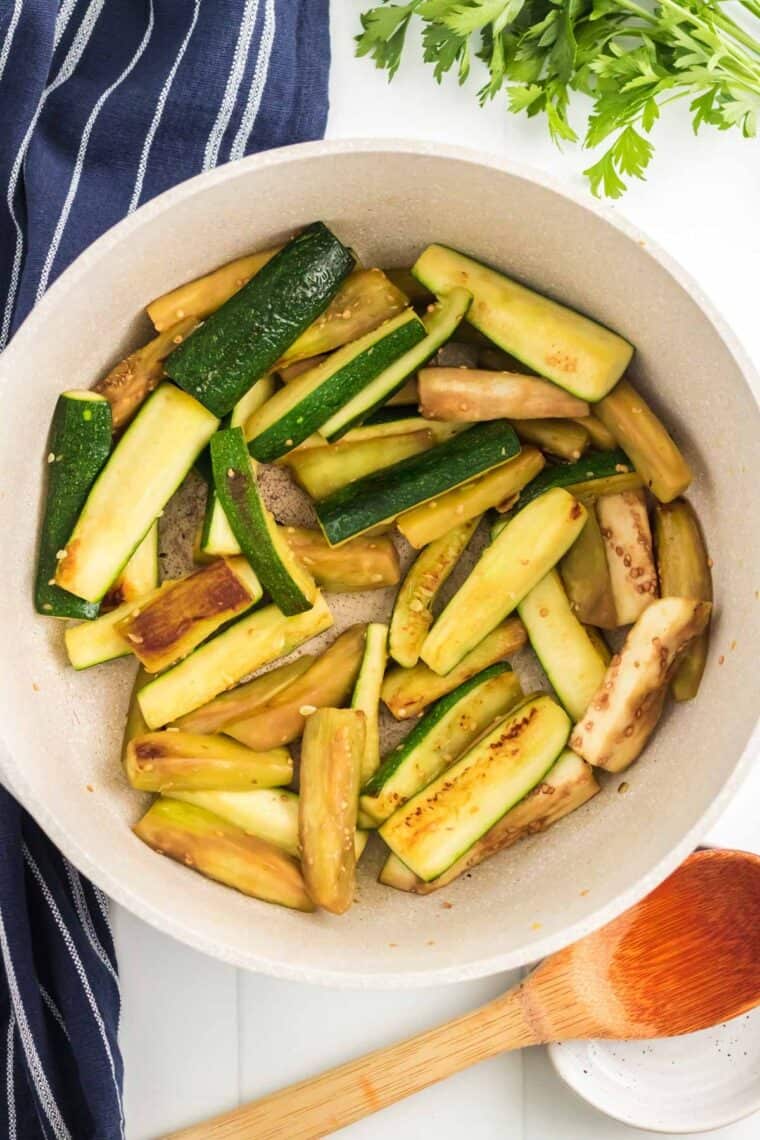 Lightly browned eggplant and zucchini in a cooking pot beside a wooden spoon