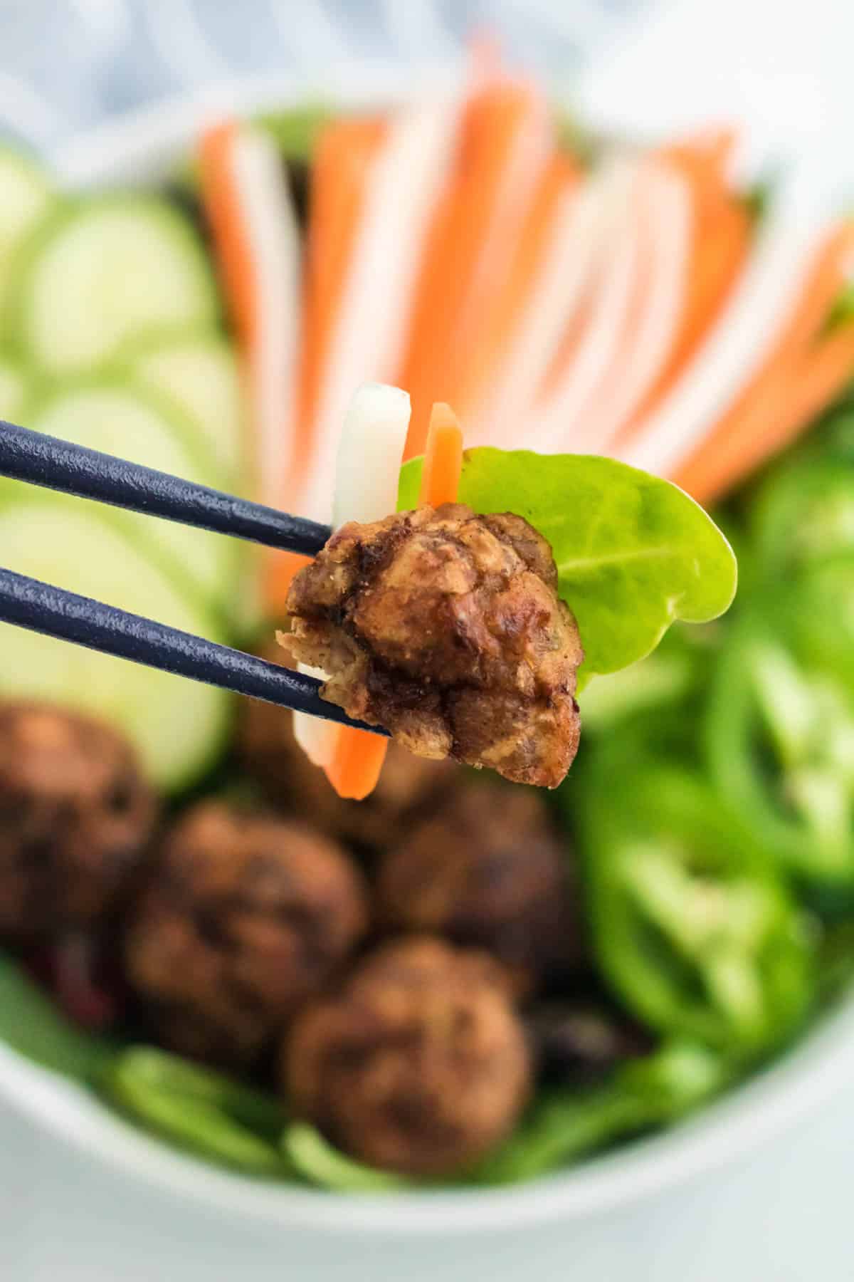 A spinach leaf, a piece of pork meatball, and a slice of pickled radish and carrot balanced between chopsticks in front of banh mi salad