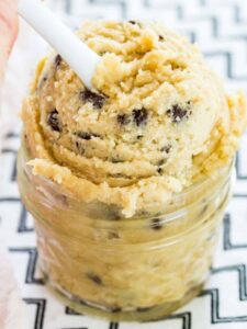 A white spoon scoops gluten-free chocolate chip cookie dough from a glass mason jar.