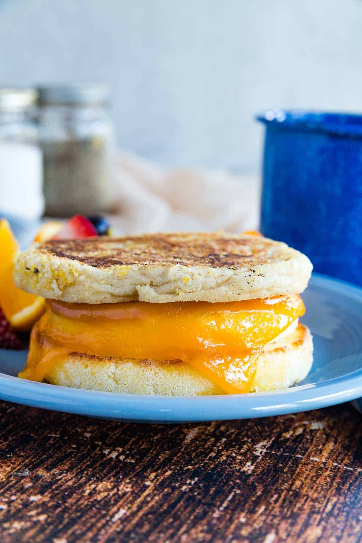 A gluten free breakfast sandwich on a blue plate next to fresh fruit, with a blue coffee mug in the background.