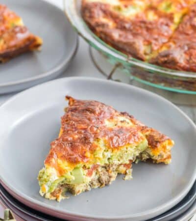 A slice of quiche on a plate with another slice and the rest of the quiche in the background.